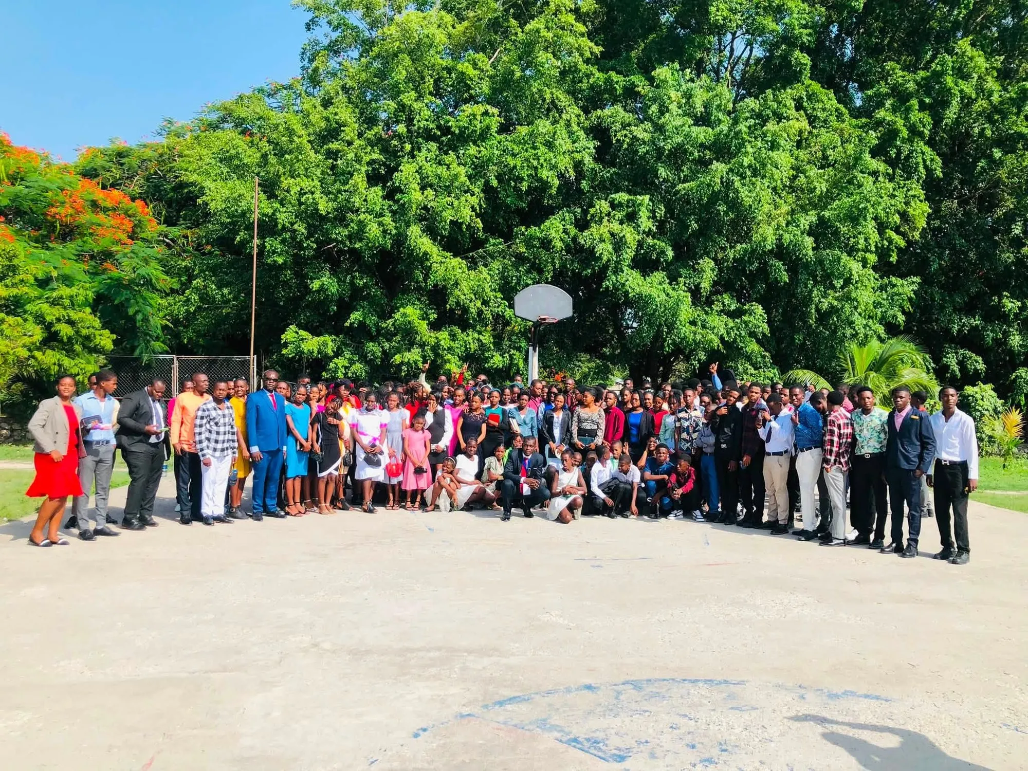 A large group of youth camp participants gathered outdoors on a court beneath tall trees