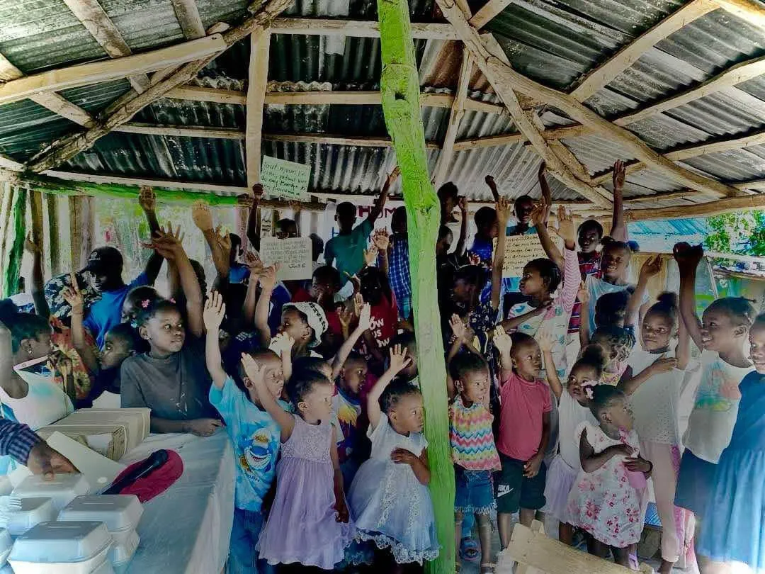 A crowd of children raising their hands under a tin-roofed shelter