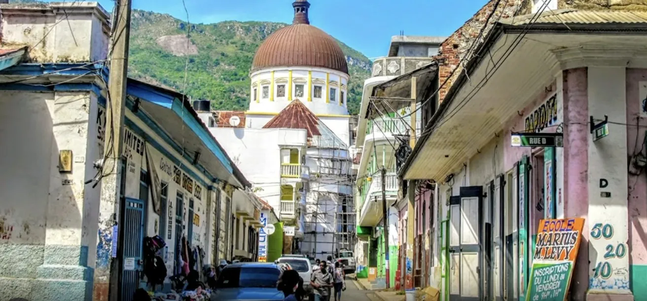 A busy street in a Haitian town with colorful buildings and a domed cathedral against the mountains