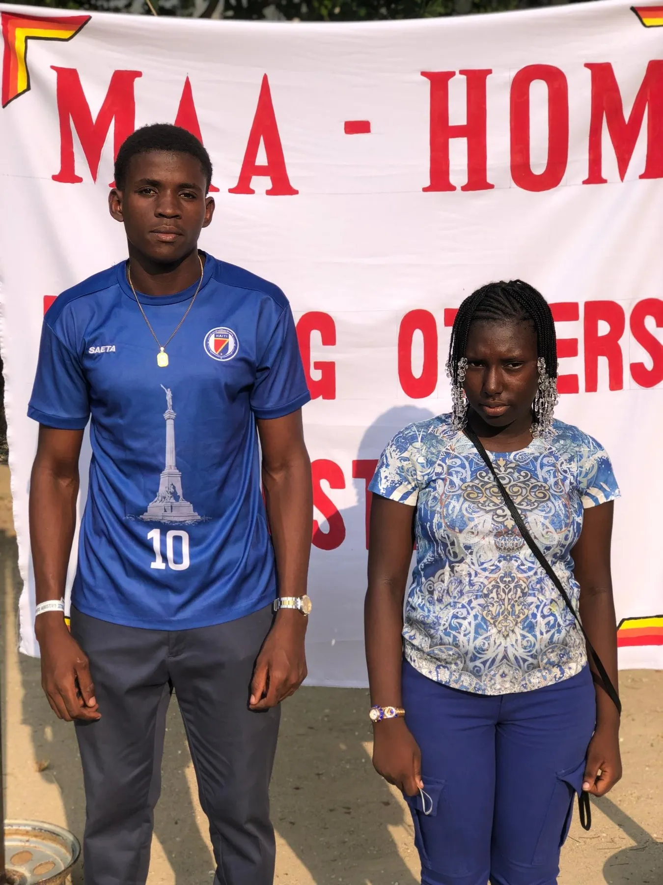 Two young people standing in front of a Helping Others Ministry banner